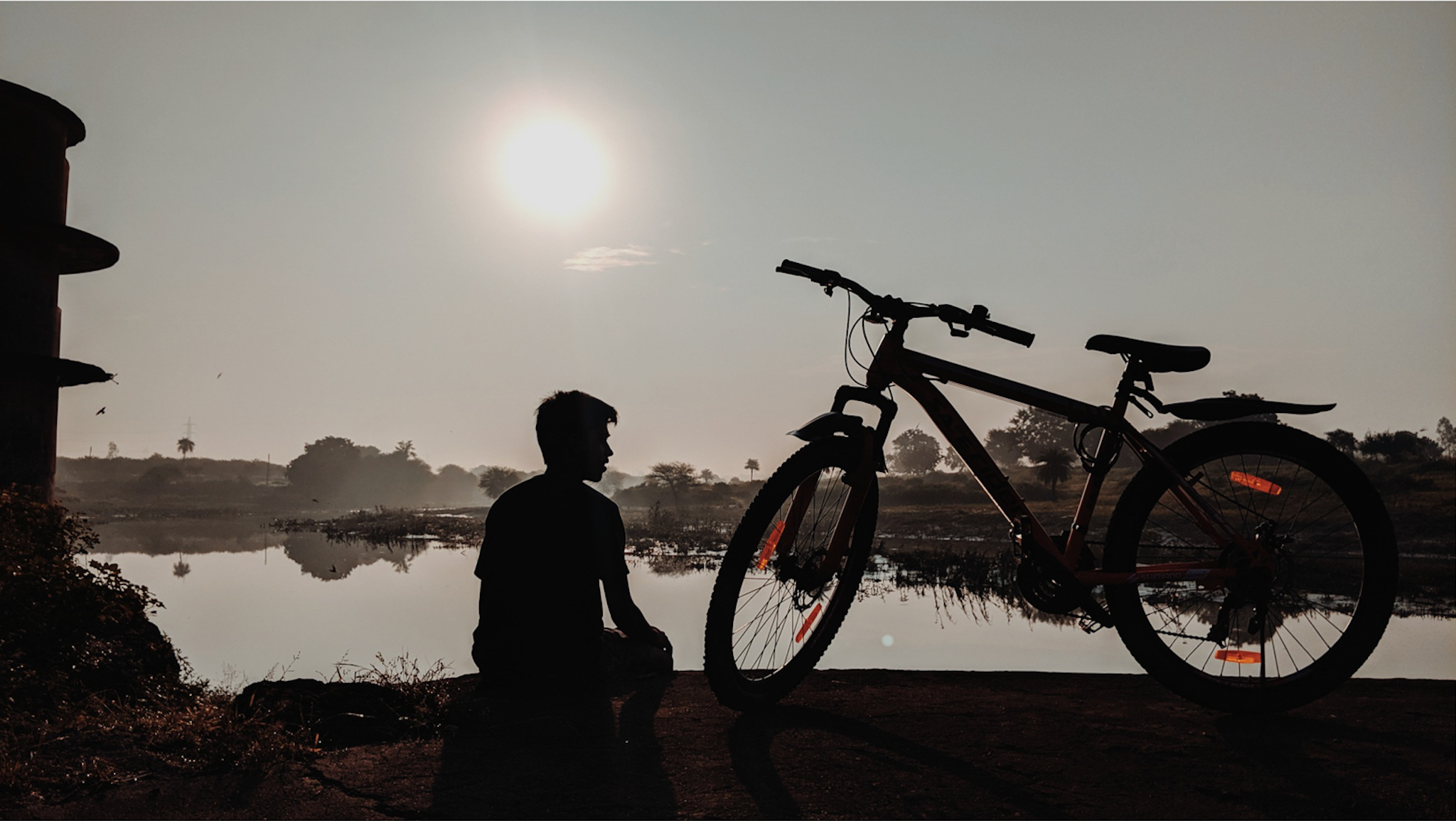 A boy sitting beside a bicycle and enjoying sunrise.